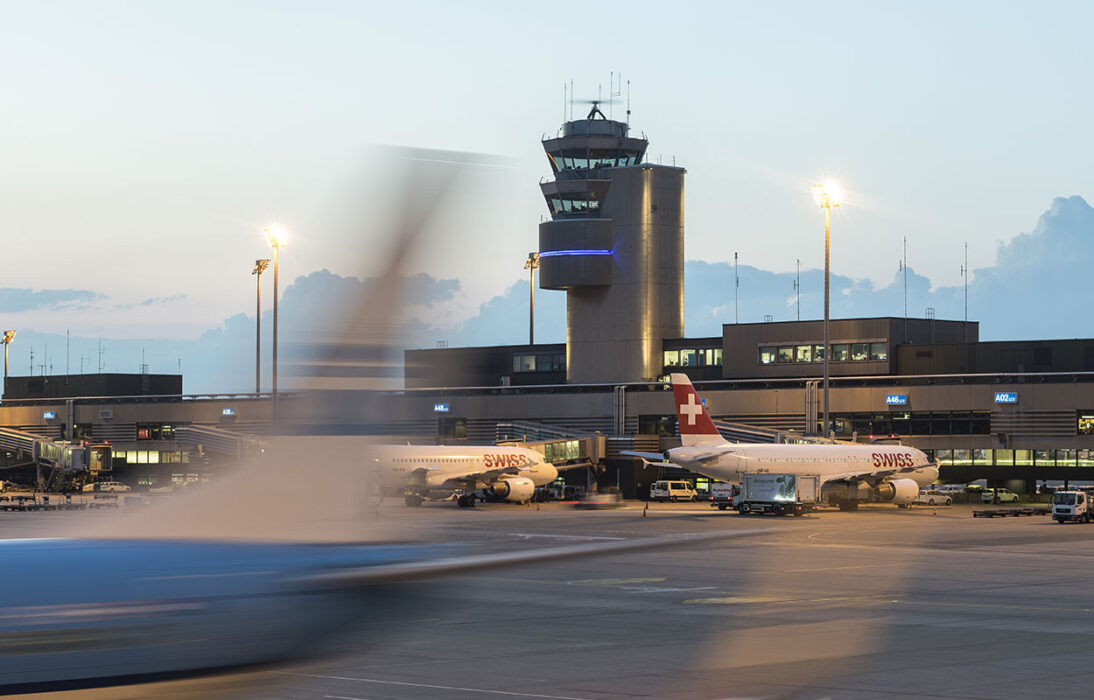 Revision_Luftfahrtgesetz_Anschluss_nicht_verlieren. Im Hintergrund ist der Tower des Flughafens Zürich erkennbar. Im Vordergrund in der Unschärfe erkennt man ein zur Startbahn rollendes Flugzeug.
